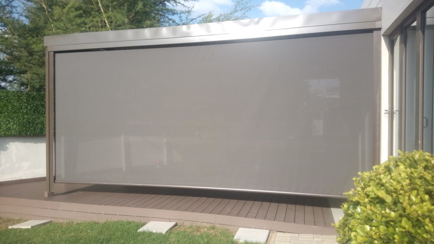 Grey fabric drop blind fully lowered on a freestanding aluminium patio structure over a composite deck