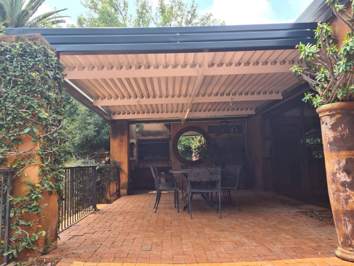 Charcoal-framed louvre awning with closed cream slats over a terracotta-paved courtyard with a built-in braai