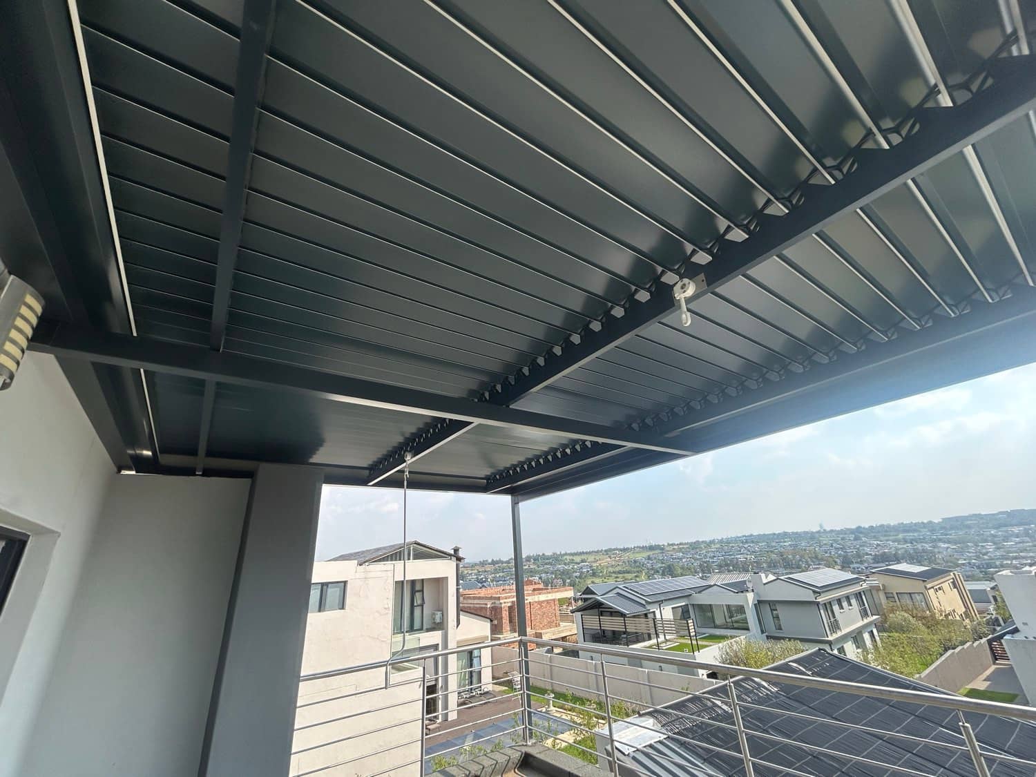 Underside of a charcoal louvre roof with slats tilted open over an upstairs balcony, rooftops and greenery in the distance