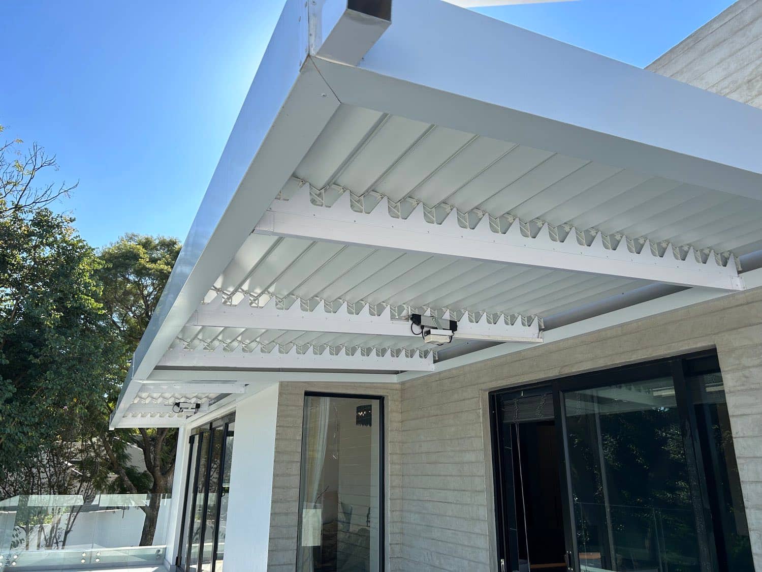 White louvre awning with motorised slats on the corner of a modern home above dark-framed glass doors