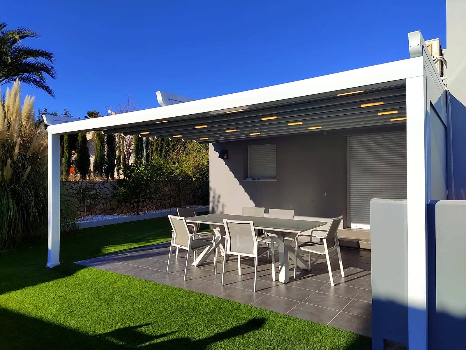 White louvre roof pergola with integrated lighting beside a lush garden