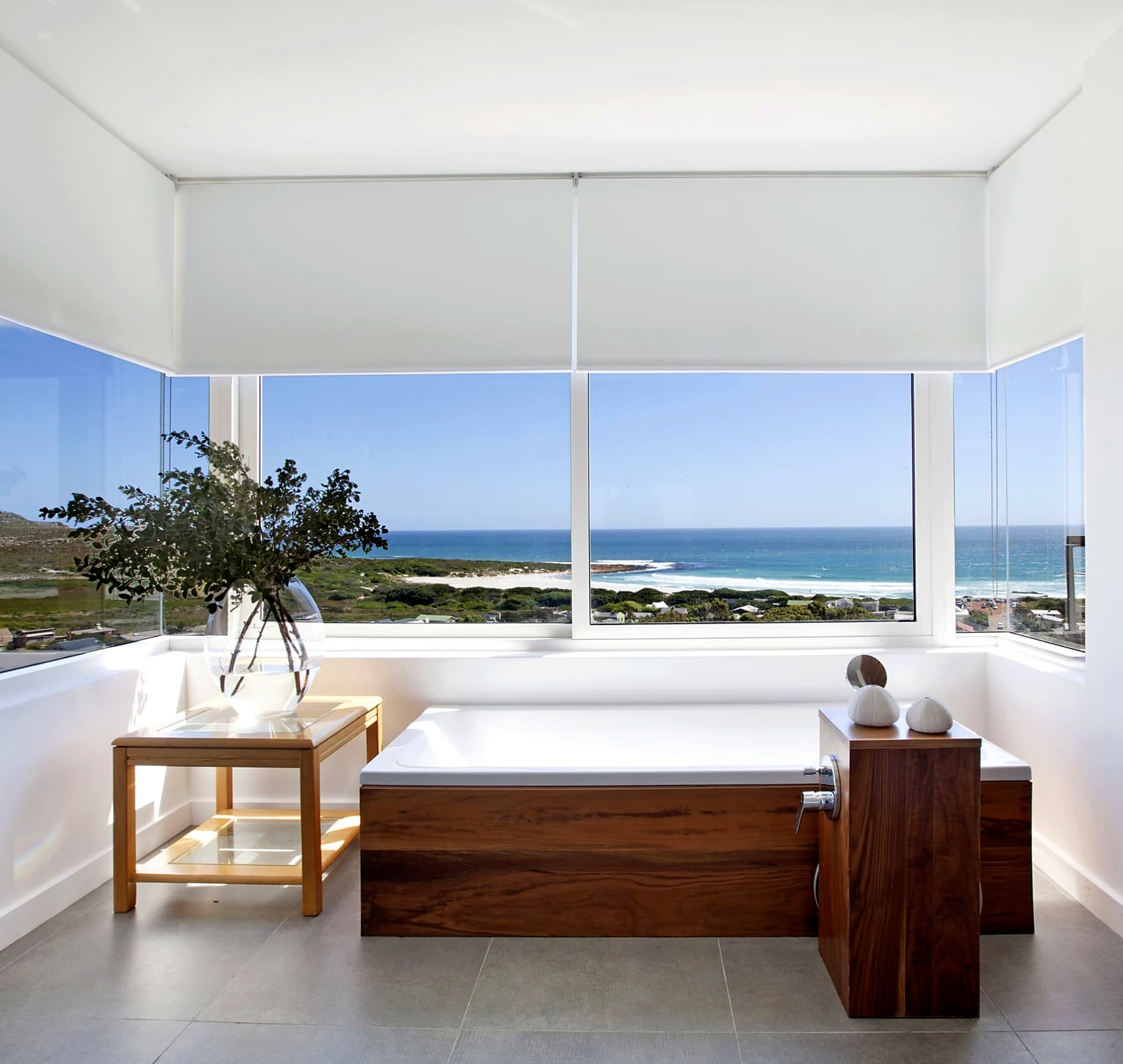 White blockout roller blinds framing an ocean view from a freestanding bath
